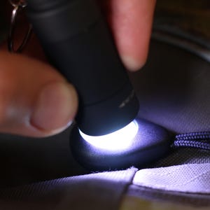 Op de afbeelding: Een zwarte zaklamp met een wit licht dat schijnt op een donkergrijs stofoppervlak.