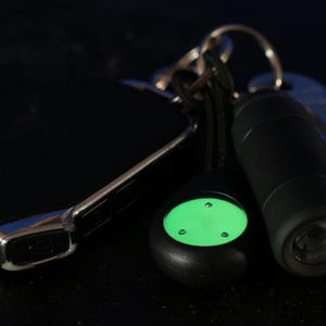 Op de afbeelding: Een zwarte sleutelhanger met een groene glow-in-the-dark knop, een zwarte zaklamp en een zilveren sleutel.