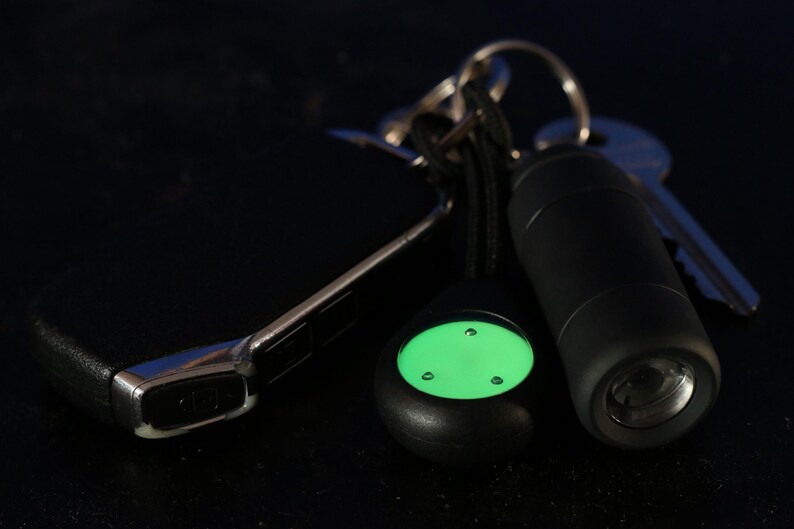 Op de afbeelding: Een zwarte sleutelhanger met een groene glow-in-the-dark knop, een zwarte zaklamp en een zilveren sleutel.