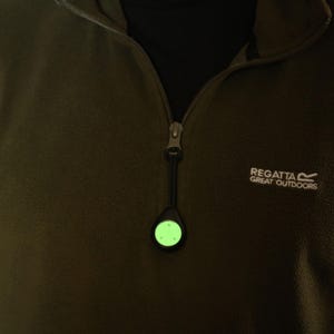 Op de afbeelding: Een groen, in het donker lichtend veiligheidslicht bevestigd aan een rits. Het licht is rond en heeft een zwarte behuizing. De rits is bevestigd aan een donkergroen fleecejack met het Regatta Great Outdoors logo.