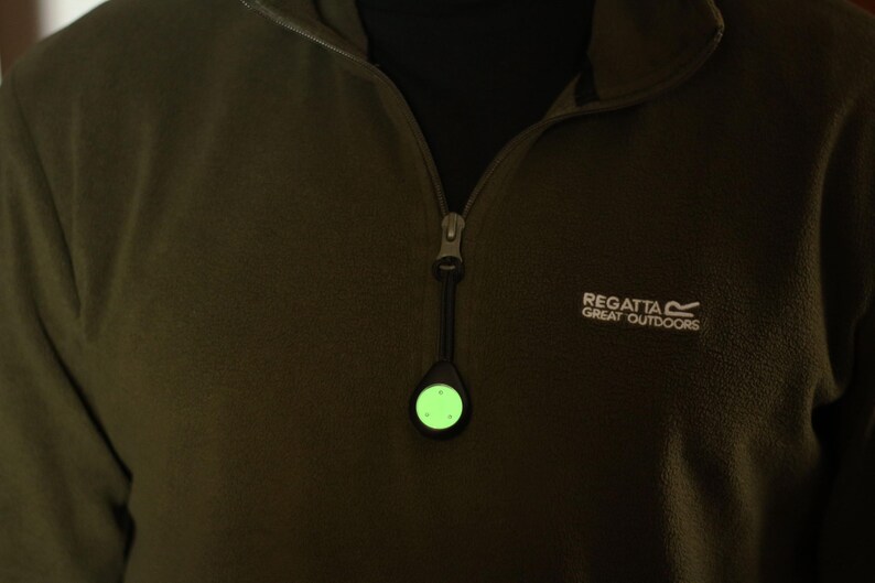 Op de afbeelding: Een groen, in het donker lichtend veiligheidslicht bevestigd aan een rits. Het licht is rond en heeft een zwarte behuizing. De rits is bevestigd aan een donkergroen fleecejack met het Regatta Great Outdoors logo.