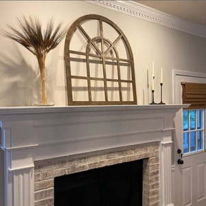 May include: A white painted brick fireplace with a white mantel and a large wooden arched window frame above it. The window frame is brown and has a grid pattern. There is a vase with dried grasses on the mantel.