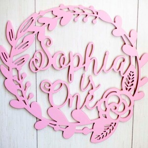 Puede incluir: Un letrero de madera rosa con un dise&ntilde;o de corona de flores. El letrero dice "Sophia is One" en una fuente cursiva.