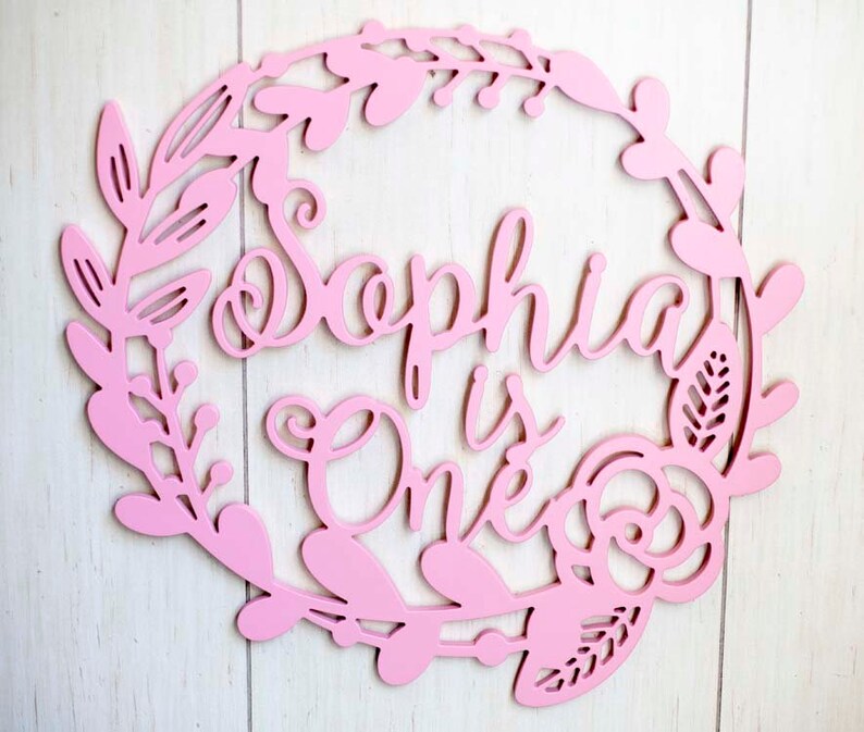 Puede incluir: Un letrero de madera rosa con un dise&ntilde;o de corona de flores. El letrero dice "Sophia is One" en una fuente cursiva.