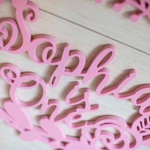 Puede incluir: Corte de madera rosa del nombre "Sophia" con la palabra "One" debajo, rodeado de un dise&ntilde;o floral.