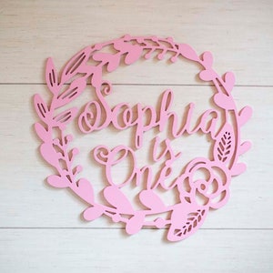 Puede incluir: Un letrero de madera rosa con un dise&ntilde;o de corona de flores. El letrero dice "Sophia is One".