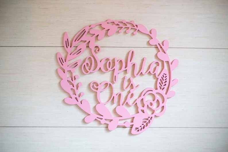 Puede incluir: Un letrero de madera rosa con un dise&ntilde;o de corona de flores. El letrero dice "Sophia is One".