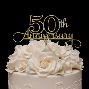 May include: Gold glitter cake topper with the text "50th Anniversary" in a cursive font. The topper is on top of a white cake decorated with white roses.