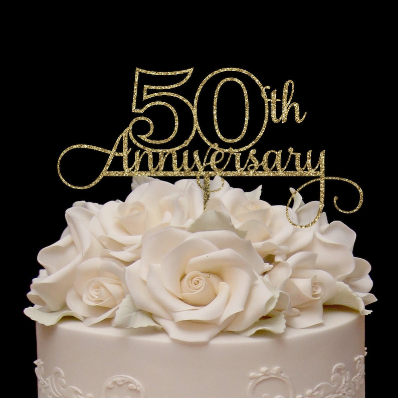 May include: Gold glitter cake topper with the text "50th Anniversary" in a cursive font. The topper is on top of a white cake decorated with white roses.