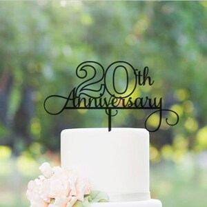 May include: Black acrylic cake topper with the text "20th Anniversary" in a decorative script font. The topper is placed on a white cake.