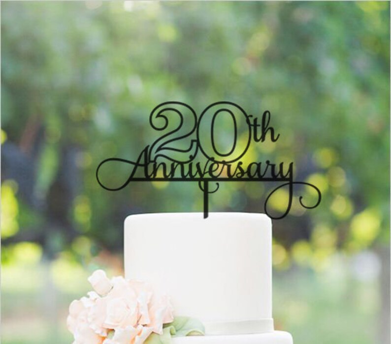 May include: Black acrylic cake topper with the text "20th Anniversary" in a decorative script font. The topper is placed on a white cake.