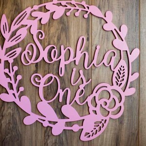 Puede incluir: Un letrero de madera rosa con un dise&ntilde;o de corona floral. El letrero dice "Sophia is One" en una fuente cursiva.