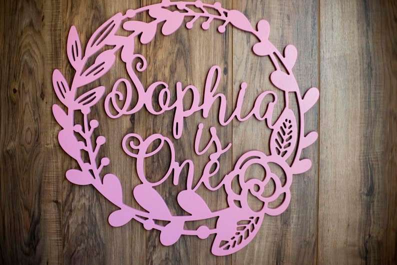 Puede incluir: Un letrero de madera rosa con un dise&ntilde;o de corona floral. El letrero dice "Sophia is One" en una fuente cursiva.