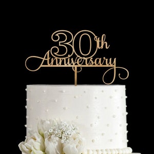 May include: A gold wooden cake topper with the text "30th Anniversary" in a cursive font. The topper is on top of a white cake with white frosting and white pearl sprinkles.