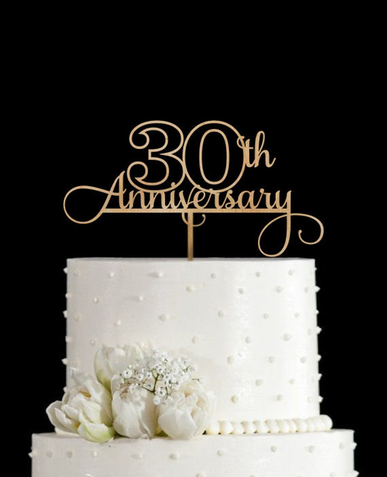 May include: A gold wooden cake topper with the text "30th Anniversary" in a cursive font. The topper is on top of a white cake with white frosting and white pearl sprinkles.