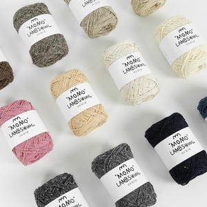 Puede incluir: Surtido de ovillos de lana de colores, incluyendo tonos de rosa, beige, gris y azul marino. Cada ovillo está etiquetado con el texto "MOMO LAMBSWOOL" en inglés y coreano. Ideal para proyectos de punto y ganchillo.