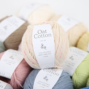 May include: Close-up of several yarn balls in various colors, including beige, pink, and blue. The central ball has a label that reads "Oat Cotton" in English and Korean, with the words "natural combed cotton collection" below. The yarn appears soft and textured.