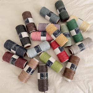 May include: A collection of colorful spools of leather cord in various shades, including green, blue, brown, pink, and yellow. Each spool has a white label with black text that reads "LEATHER EDITION". The spools are arranged on a white surface.