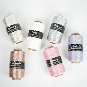 May include: An assortment of metallic cord cones in various colors, including silver, white, pink, lavender, and bronze. Each cone has a black label with the word "Galaxy" and "Metallic Cord Cone" printed on it. The cones are arranged on a white surface.