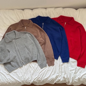 May include: Four zip-up knit sweaters in gray, brown, navy blue, and red. Each sweater has a collared neckline and a silver zipper. The sweaters are displayed on a white, textured surface.