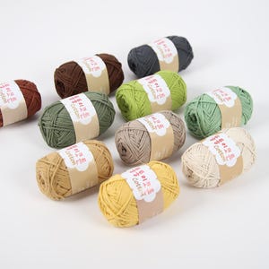 May include: A collection of colorful yarn skeins in various shades, including brown, green, yellow, and gray. Each skein is wrapped with a paper band featuring text. These cotton yarn balls are ideal for knitting and crochet projects.
