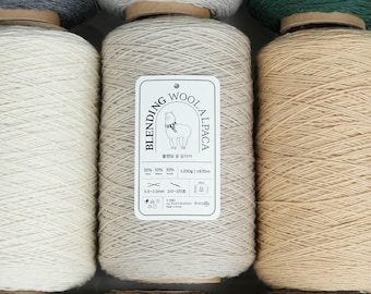 Pode incluir: Close-up de vários rolos de fio em várias cores, incluindo branco, cinza e bege. Um rótulo em um rolo diz "Blending Wool Alpaca" com detalhes sobre a composição da fibra. O fio é enrolado em rolos de madeira.