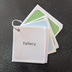 Op de afbeelding: Een stapel vierkante flashcards die bij elkaar worden gehouden door een zilveren metalen ring. De bovenste kaart is lichtgrijs met het woord Fallacy in het zwart gedrukt. Daaronder liggen extra kaarten in lichtblauw en groen met afgeronde hoeken.