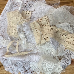 May include: A collection of white and cream lace trims, including a variety of patterns and widths. The lace is arranged in a pile on a wooden surface.