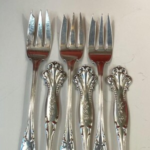 May include: Six silver forks with ornate handles. The forks have three tines and a decorative design on the handle. The handles have a floral pattern and a curved shape. The forks are arranged on a light-colored surface.