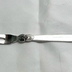 May include: A silver-colored, two-pronged fork with a decorative floral design on the handle. The fork has a pointed tip on each prong. The handle is long and slender, and the fork is likely made of stainless steel.