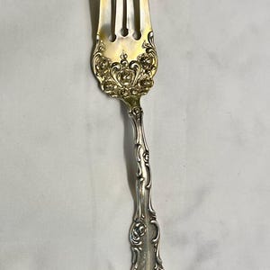 May include: A silver fork with four tines and a decorative handle. The fork's tines are gold-colored, and the handle features ornate floral detailing. The silverware is likely vintage or antique.