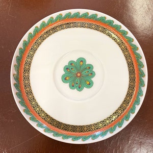 May include: White ceramic saucer with a decorative design. The rim features a green and orange leaf pattern, with a gold and black geometric border. The center has a green and blue floral design. A stylish addition to any table setting.