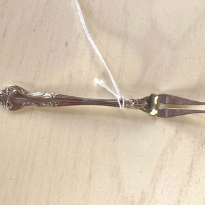 May include: A silver-colored, ornate serving fork with two tines. The handle features a decorative floral design. The fork is lying on a light-colored wooden surface.