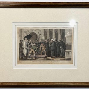 May include: A framed historical print titled "OFFENDER AT THE FUNERAL OF HIS WIFE" featuring a monochromatic scene of a group of people gathered outside a stone church. The artwork is encased in a gold-toned wooden frame with a cream-colored mat.