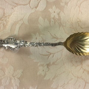 May include: An antique silver spoon with a gold-colored bowl. The spoon features ornate detailing on the handle and a scalloped bowl design. The spoon is likely used for serving or decorative purposes.