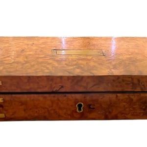 May include: A rectangular wooden box with a polished burl wood grain finish. The box features brass corner accents on the front and sides, a central brass keyhole escutcheon, and a rectangular brass nameplate centered on the top lid surface.