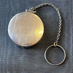 May include: A round, silver-toned compact with a patterned surface and a chain attached to a ring. The compact is closed and has a hinged lid. The chain and ring are also silver-toned.