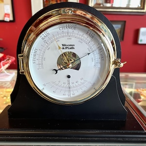 Puede incluir: Un barómetro de madera negra con un bisel dorado y una esfera blanca. La esfera muestra indicadores meteorológicos y lecturas de temperatura. El barómetro está etiquetado como "Weems & Plath".