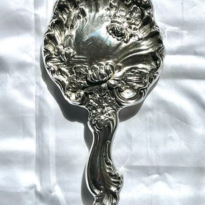 May include: A silver-toned hand mirror with an ornate floral design. The mirror's surface reflects light, and the handle is curved with detailed embellishments. The overall design is vintage and elegant, with a classic aesthetic.