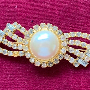 May include: A gold-tone brooch with a large faux pearl center surrounded by clear rhinestones. The brooch is shaped like a bow with three rows of rhinestones on each side.