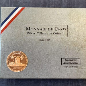 Puede incluir: Una caja gris con una franja roja, blanca y azul en la parte superior. La caja tiene el texto "MONNAIE DE PARIS" y "Pièces "Fleurs de Coins"" en negro. La caja también tiene el texto "Série 1969" en negro. La caja tiene una moneda de oro con el texto "DANT PRET" y "MONNGTEURS DE LA MONNTE DE PARIS ENG" en el centro. La caja también tiene una etiqueta blanca con el texto "Exemplaires Numismatiques" y "MADE IN FRANCE" en negro.