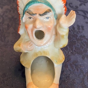 May include: A vintage ceramic figurine of a Native American chief with an open mouth and raised hand. The figurine features a headdress with orange and white feathers, a green headband, and a tan-colored outfit. The figure has an open hole in the center.