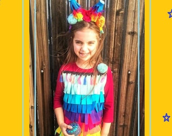 May include: A child wearing a colourful pinata costume with a rainbow headband. The costume is made of paper and has fringe around the bottom. The child is holding a small, sparkly ball.