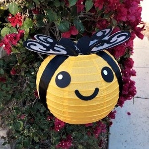 May include: A yellow paper lantern shaped like a bee with black stripes, wings, and a smiling face. The bee lantern is hanging from a branch of a bush with pink flowers.