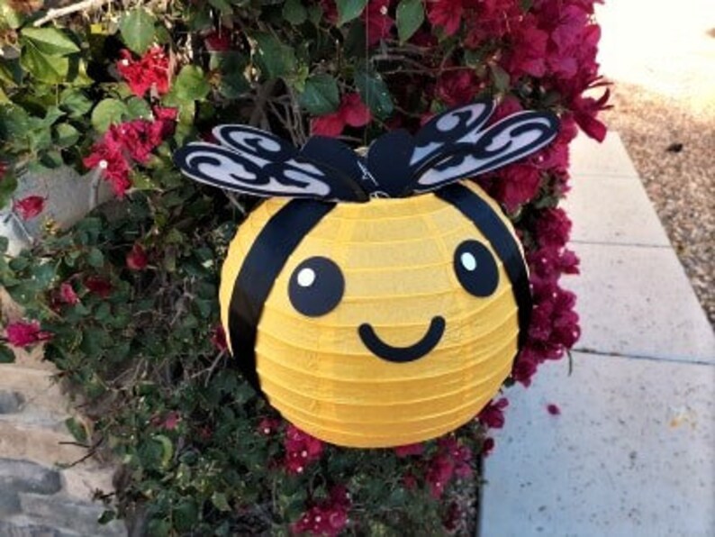May include: A yellow paper lantern shaped like a bee with black stripes, wings, and a smiling face. The bee lantern is hanging from a branch of a bush with pink flowers.