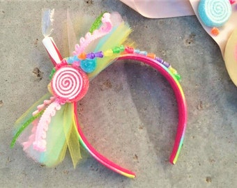 May include: A pink headband with a rainbow stripe, decorated with colorful candy and tulle. The headband is adorned with a large swirl lollipop, beads, and a green streamer.