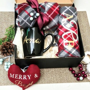 May include: A black gift box containing a red and grey plaid scarf, a black mug with the name "Anna" on it, and a red heart-shaped ornament that says "Merry & Bright".