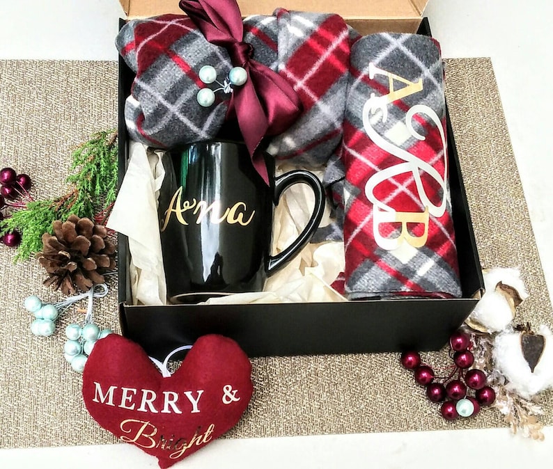 May include: A black gift box containing a red and grey plaid scarf, a black mug with the name "Anna" on it, and a red heart-shaped ornament that says "Merry & Bright".