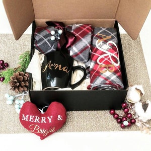 May include: A black gift box with a red and gray plaid blanket, a black mug with the name "Anna" in gold lettering, and a red heart-shaped pillow with the text "Merry & Bright" in gold lettering.