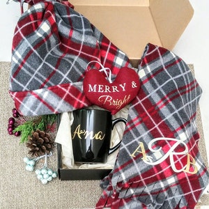 May include: A gift box filled with a red and grey plaid hat, scarf, and mug. The mug is black with the name "Ana" written in gold lettering. A red heart with the words "Merry & Bright" is included in the box.